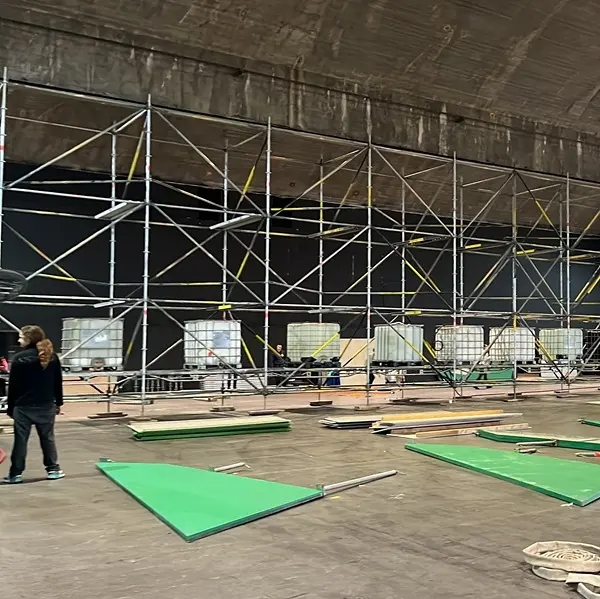 Een grote binnenruimte met steigers, groene driehoekige panelen en diverse bouwmaterialen op de vloer. Er zijn verschillende mensen aanwezig die bezig lijken te zijn met het opbouwen of afbreken van een constructie of podium.